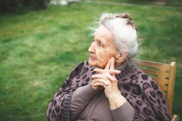 Elderly woman 80-90 years with gray hair sitting on a wooden chair in a garden with her hands clasped, thoughtfully looking away, concept of aging and contemplation