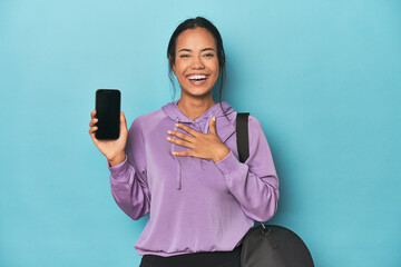 Filipina ready for gym with phone on blue laughs out loudly keeping hand on chest.