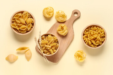 Composition with bowls of different types of raw pasta and wooden cutting board on beige background