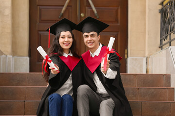 Couple of happy graduating students with diplomas sitting on stairs near university
