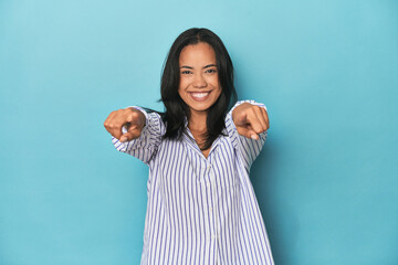 Filipina young woman on blue studio cheerful smiles pointing to front.