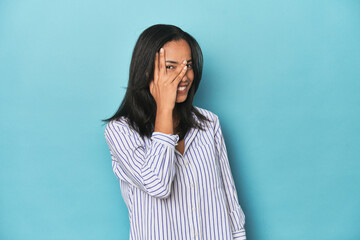 Filipina young woman on blue studio blink at the camera through fingers, embarrassed covering face.