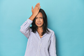 Filipina young woman on blue studio forgetting something, slapping forehead with palm and closing...
