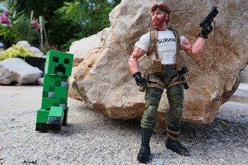 Naklejka premium GI JOE action figure of survival instructor Outback, verion 2023 Classified edition, armed with pistol, ready to attack explosive green LEGO Minecraft Creeper from behind a shoreline rock.