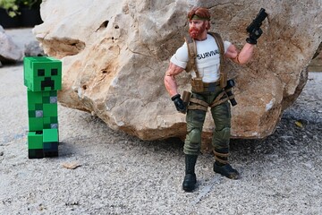Naklejka premium GI JOE action figure of survival instructor Outback, verion 2023 Classified edition, armed with pistol, ready to surprise explosive green LEGO Minecraft Creeper from behind a shoreline rock.