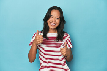 Filipina young woman on blue studio raising both thumbs up, smiling and confident.