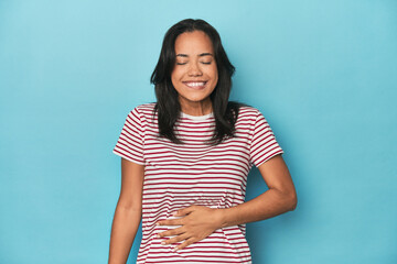 Filipina young woman on blue studio touches tummy, smiles gently, eating and satisfaction concept.