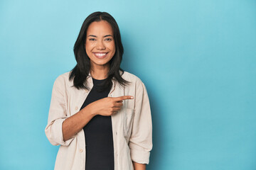 Filipina young woman on blue studio smiling and pointing aside, showing something at blank space.