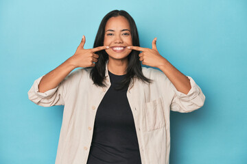 Filipina young woman on blue studio smiles, pointing fingers at mouth.