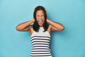 Filipina young woman on blue studio covering ears with hands trying not to hear too loud sound.