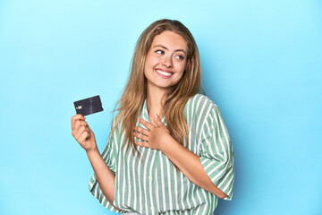 Young blonde with a credit card in blue studio laughs out loudly keeping hand on chest.
