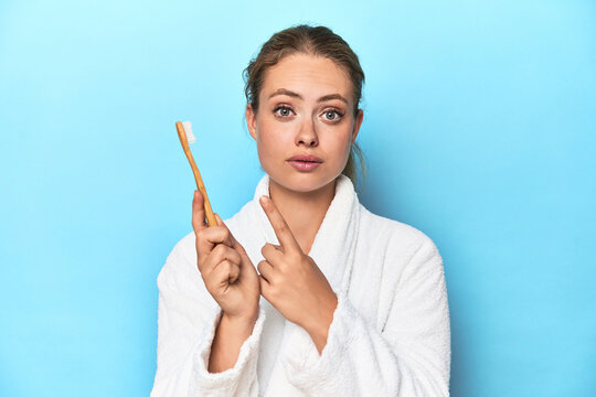 Blonde in bathrobe with a toothbrush in blue studio pointing to the side - Powered by Adobe