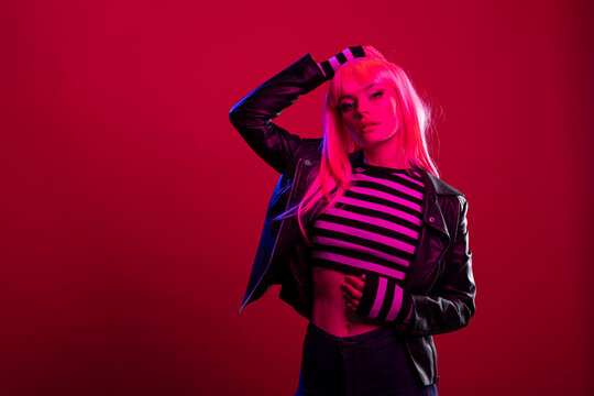 Stylish young woman with long hair posing under dramatic pink and blue lighting. Beautiful female model wearing leather jacket and striped crop top, exudes bold, edgy fashion and urban attitude.