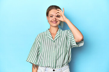 Blonde young woman in green striped shirt on blue background excited keeping ok gesture on eye.