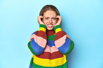 Blonde young woman in multicolor sweatshirt on blue background covering ears with hands.
