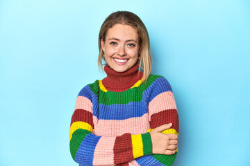 Blonde young woman in multicolor sweatshirt on blue background who feels confident, crossing arms...