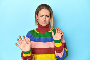 Blonde young woman in multicolor sweatshirt on blue background rejecting someone showing a gesture...