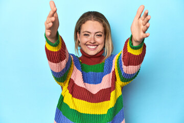 Blonde young woman in multicolor sweatshirt on blue background feels confident giving a hug to the...