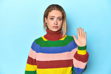 Blonde young woman in multicolor sweatshirt on blue background smiling cheerful showing number five...