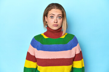 Blonde young woman in multicolor sweatshirt on blue background blows cheeks, has tired expression....