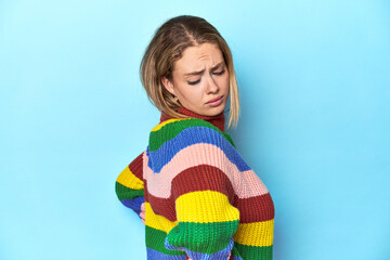 Blonde young woman in multicolor sweatshirt on blue background suffering a back pain.