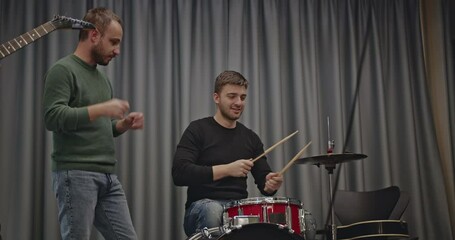 A music band practices before a concert. The drummer plays while the guitarist looks on, giving feedback and suggestions.