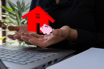 Close-up of a businesswoman's hands holding a house icon with a piggy bank, sitting at a desk in...