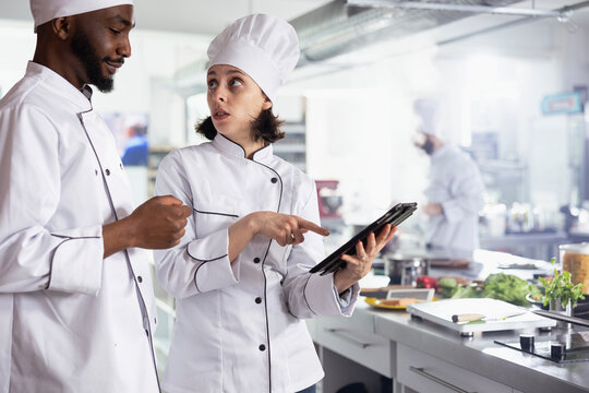 In a restaurant kitchen, diverse chefs in uniform using a tablet to find new cooking combinations ideas. Cooking professionals doing food prep with raw materials and other ingredients, fine dining.