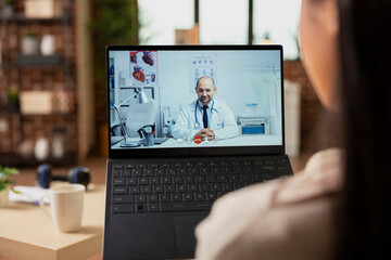 Back view of woman making video call with caucasian male doctor from home. Close up of female patient using laptop for remote consultation with general practitioner through telehealth service.
