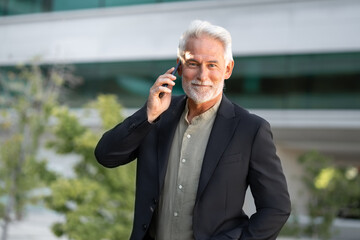 Senior businessman in suit smiling and holding smartphone during call outside modern office. Bright...