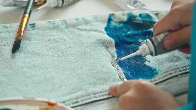 A close-up shot of a child customizing their clothes by applying paint to their jeans and creating a unique design. High angle.