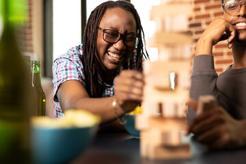 Happy black woman filled with laughter, pulling block during wooden tower game, as male friend watches nearby with a smile. Game day excitement builds during their cozy home gathering.