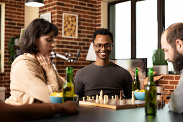 Cheerful black man wearing glasses, seated at board game session with caucasian friends. African American guy smiling playfully at camera during casual chess match in cozy brick wall room.