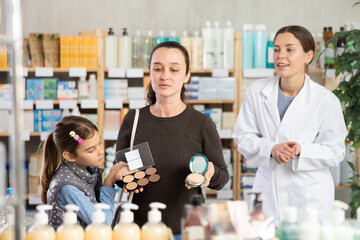 Woman of European appearance chooses decorative cosmetics in a pharmacy against the background of a pharmacist. Buyer, together with daughter, chooses powder for every day in the pharmacy