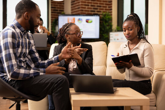 African American startup team gathers around, reviewing documents in modern workspace. Professional black woman explains marketing strategies on clipboard during business meeting.