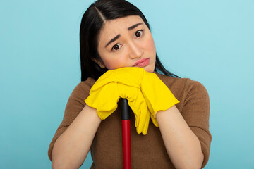 Frustrated asian woman in yellow rubber gloves leans on mop, showing displeasure and signs of...