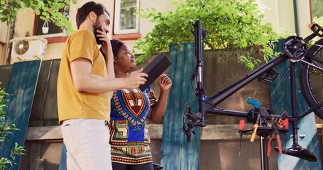 Youthful multiethnic couple utilizing digital device for repairing and maintaining modern bicycle. In home yard vibrant man and sporty black woman exploring the internet for bike maintenance.