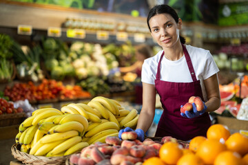 Adult woman seller in apron puts fresh peaches on display in vegetable shop