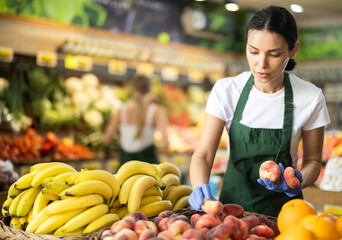 Saleswoman in an apron stands near the counter and puts ripe flat peaches next to bananas. Process of displaying fruits in a supermarket