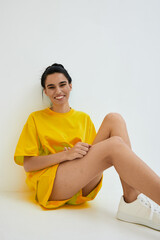 Smiling woman in yellow casual outfit sitting against white wall, showing relaxed mood, stylish...
