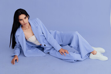 Young woman reclining on purple floor wearing pastel blue suit and white sneakers, dark hair,...