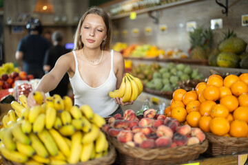 Young woman buyer choosing bunch of fresh bananas in vegetable shop
