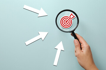 Woman holding magnifying glass with target inside against light blue background, closeup. Arrows...
