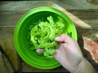 close-up photo of a woman's hand picking up slices of raw bitter melon in a basin on a wooden...