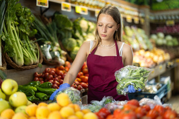 Young woman seller in apron puts green onions on display in vegetable shop