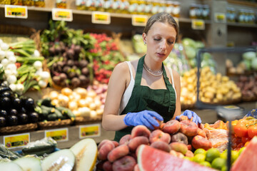 Young woman seller in apron puts fresh flat peaches on display in vegetable shop