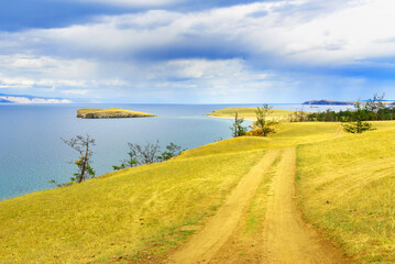 Lake Baikal stretches under a soft blue sky. Calm waters meet green hills and sandy beaches,...