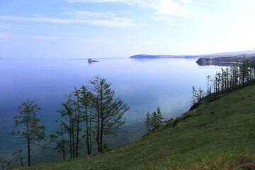 Lake Baikal stretches under a soft blue sky. Calm waters meet green hills and sandy beaches, creating a breathtaking natural scene
