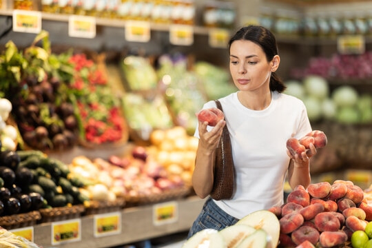 Woman near vegetable stand in store choose flat peach. Client view ripeness of fruit, take out fruits from showcase, collects grocery basket. Girl pull out ripest fruits from large pile of fruits.