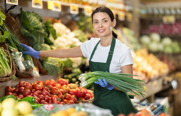 Girl puts ripe bunch of green onion in box on display case, arranges assortment in vegetable shop. Worker puts vegetables in pile pyramid. Employee reorganizes showcase ..